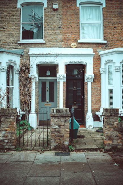 A row of white-painted terraced houses on Mill Lane in West Hampstead, each featuring dark blue garage doors and wooden external stairs leading to upper-level entrances. The windows are framed in whit